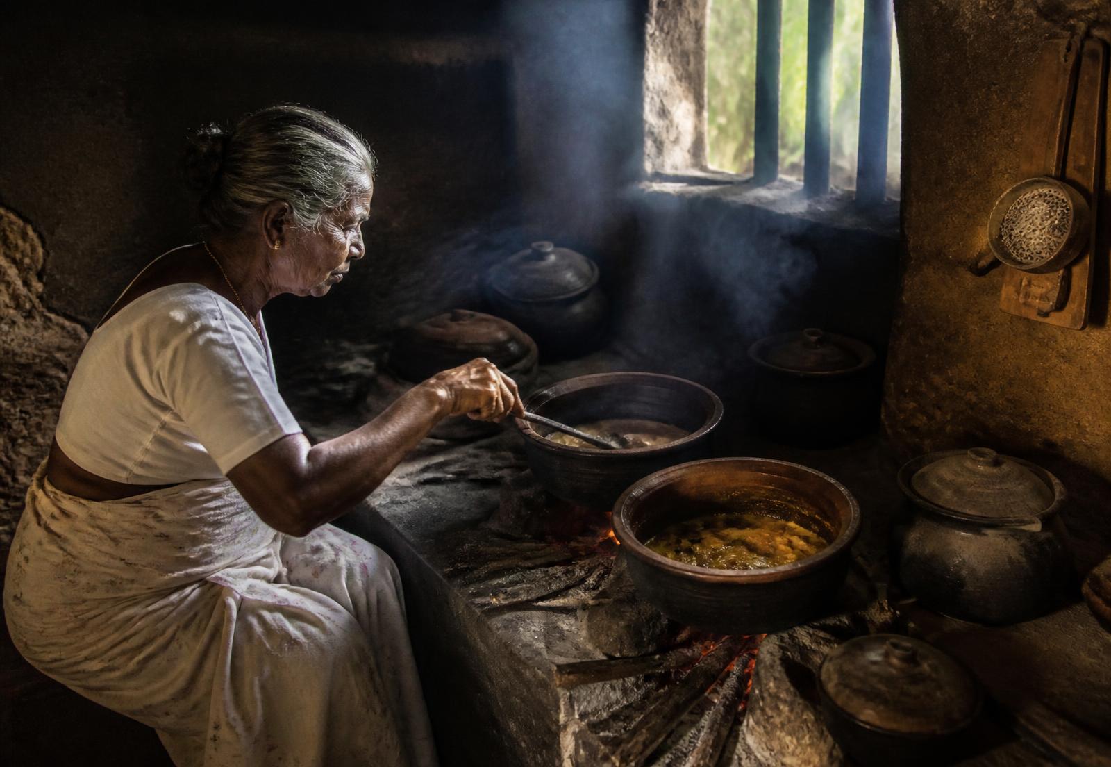 An elder cooking curry in clay pots over a wood hearth in a Sri Lankan village kitchen