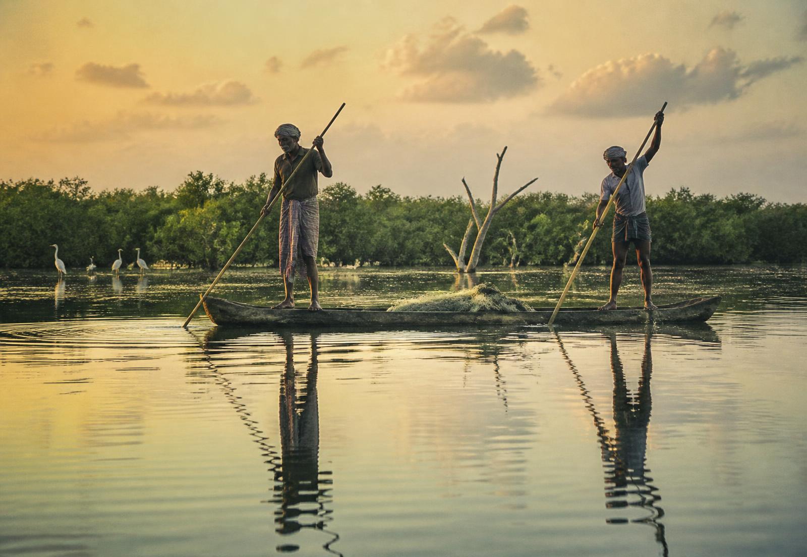 Two fishermen poling a dugout canoe across a mangrove lagoon at golden hour